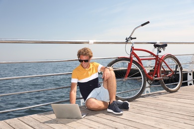 Attractive man with laptop and bike near sea on sunny day