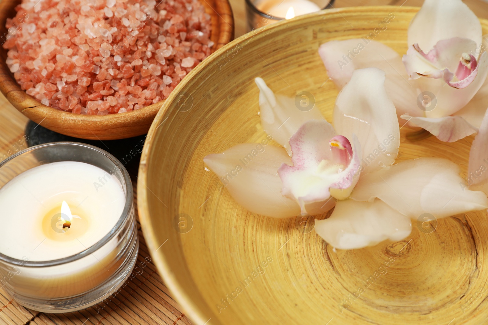 Photo of Bowl with water, flowers, sea salt and burning candles on bamboo mat, closeup. Spa treatment