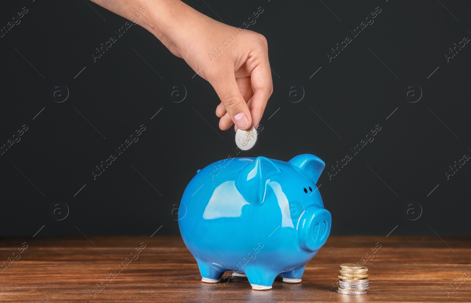 Photo of Woman putting coin into piggy bank on wooden table against dark background. Pension planning