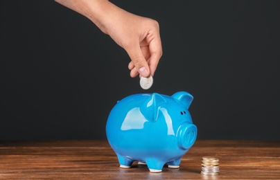 Woman putting coin into piggy bank on wooden table against dark background. Pension planning