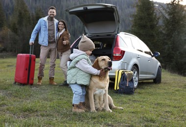 Photo of Dog, cute little girl and her parents near car in mountains. Family traveling with pet