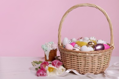 Photo of Wicker basket with festively decorated Easter eggs and beautiful tulips on white marble table against pink background. Space for text