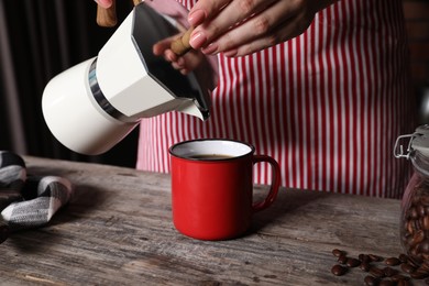 Photo of Woman pouring aromatic coffee from moka pot into cup at wooden table indoors, closeup