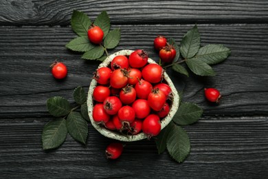 Photo of Ripe rose hip berries with green leaves on black wooden table, flat lay