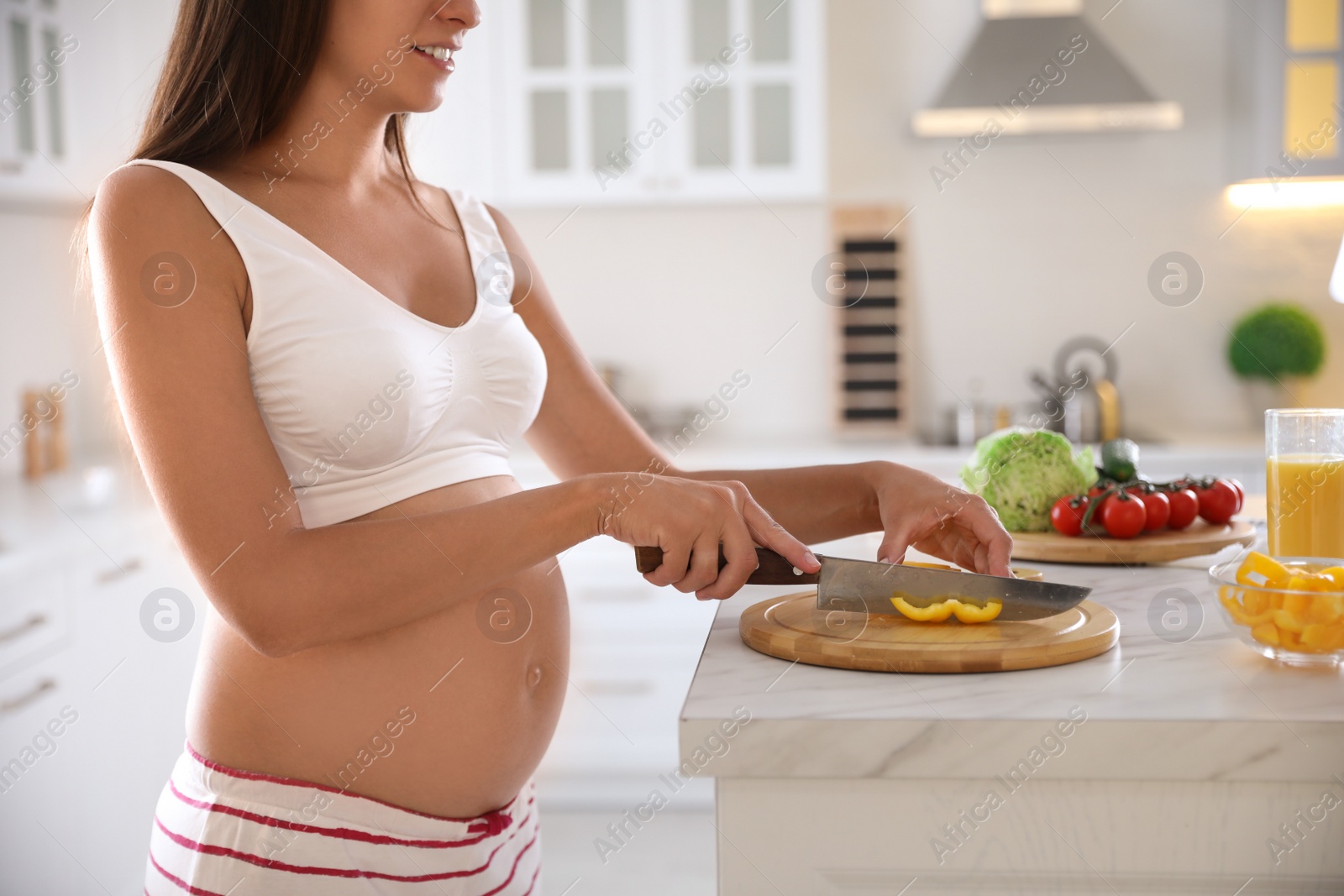 Photo of Young pregnant woman cutting yellow bell pepper at table in kitchen, closeup. Taking care of baby health