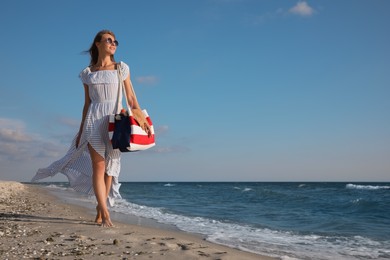 Beautiful woman with beach bag walking on sand near sea