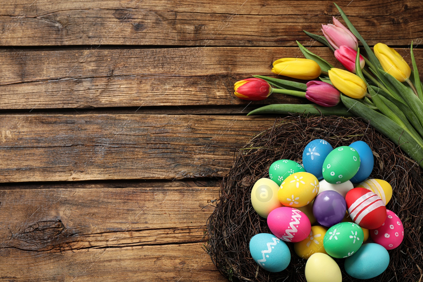 Photo of Colorful Easter eggs in decorative nest and tulips on wooden background, flat lay. Space for text