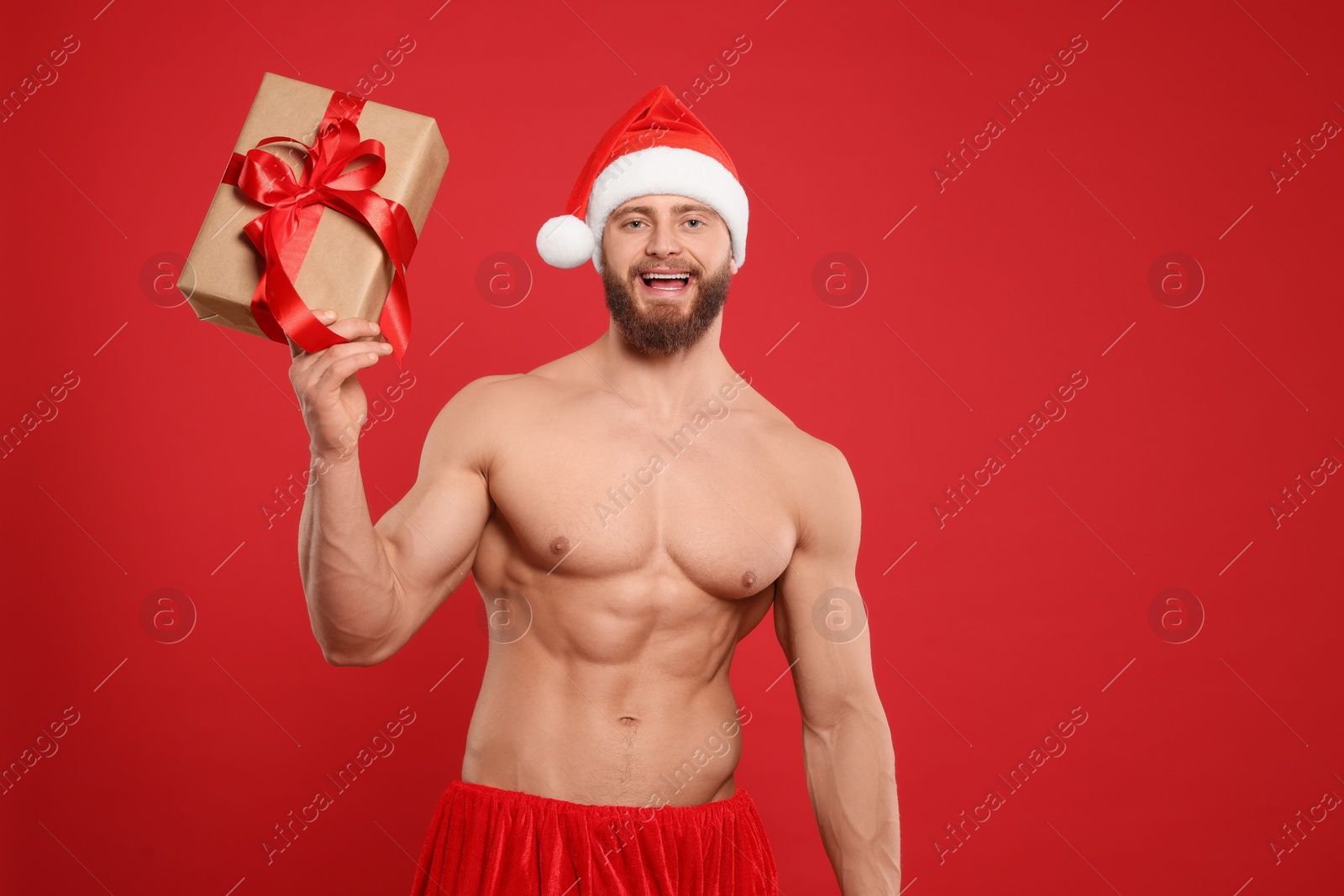 Photo of Attractive young man with muscular body in Santa hat holding Christmas gift box on red background