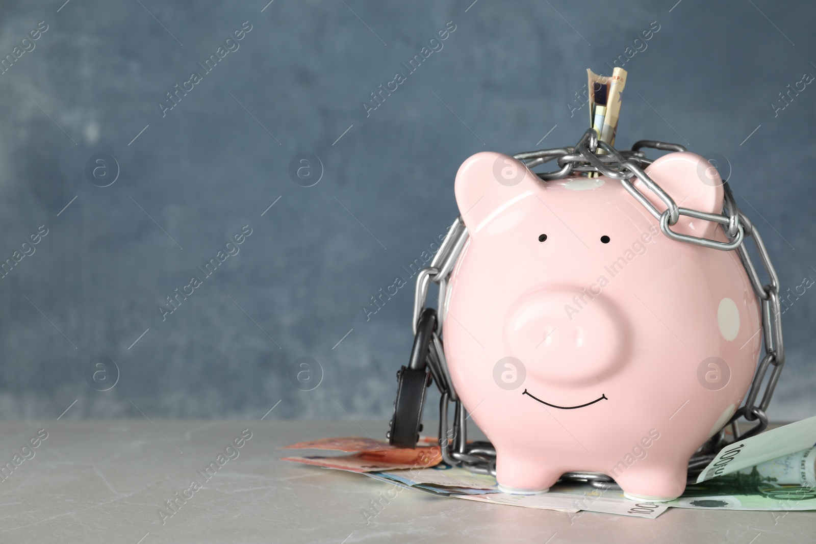 Photo of Piggy bank with steel chain, padlock and banknotes on grey marble table, space for text. Money safety concept