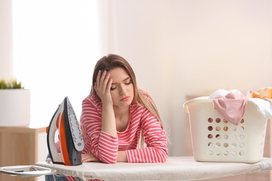 Photo of Young tired woman with iron and basket of clean laundry at board indoors