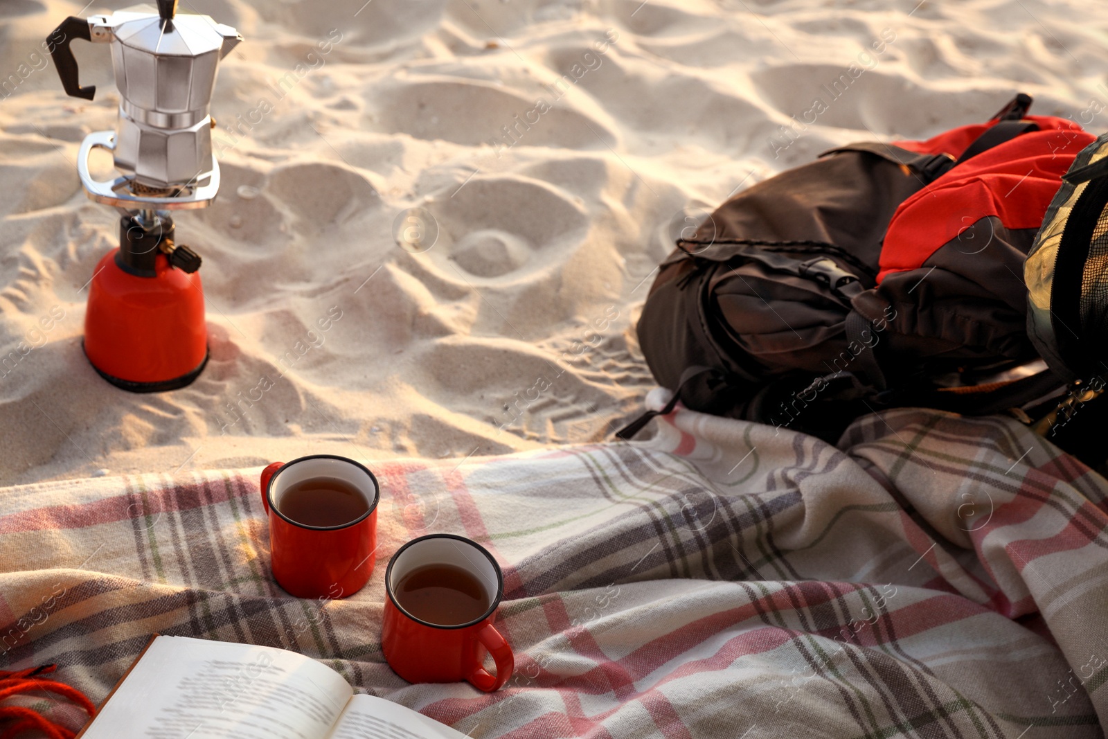 Photo of Cups of tea, book on blanket near camping stove with moka pot and backpack at sandy beach
