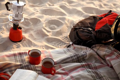 Cups of tea, book on blanket near camping stove with moka pot and backpack at sandy beach