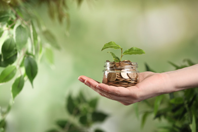 Woman holding coins and green sprout on blurred background, closeup. Money savings