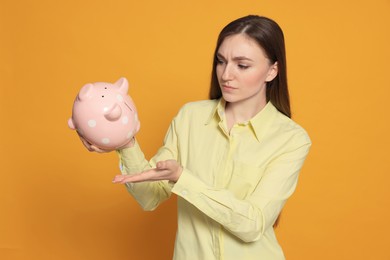 Sad young woman with piggy bank on orange background