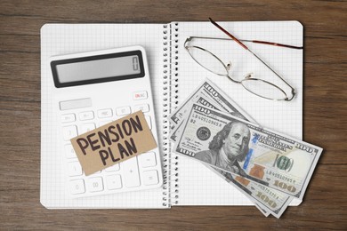 Photo of Card with phrase Pension Plan, calculator, dollar banknotes, notebook and glasses on wooden table, top view