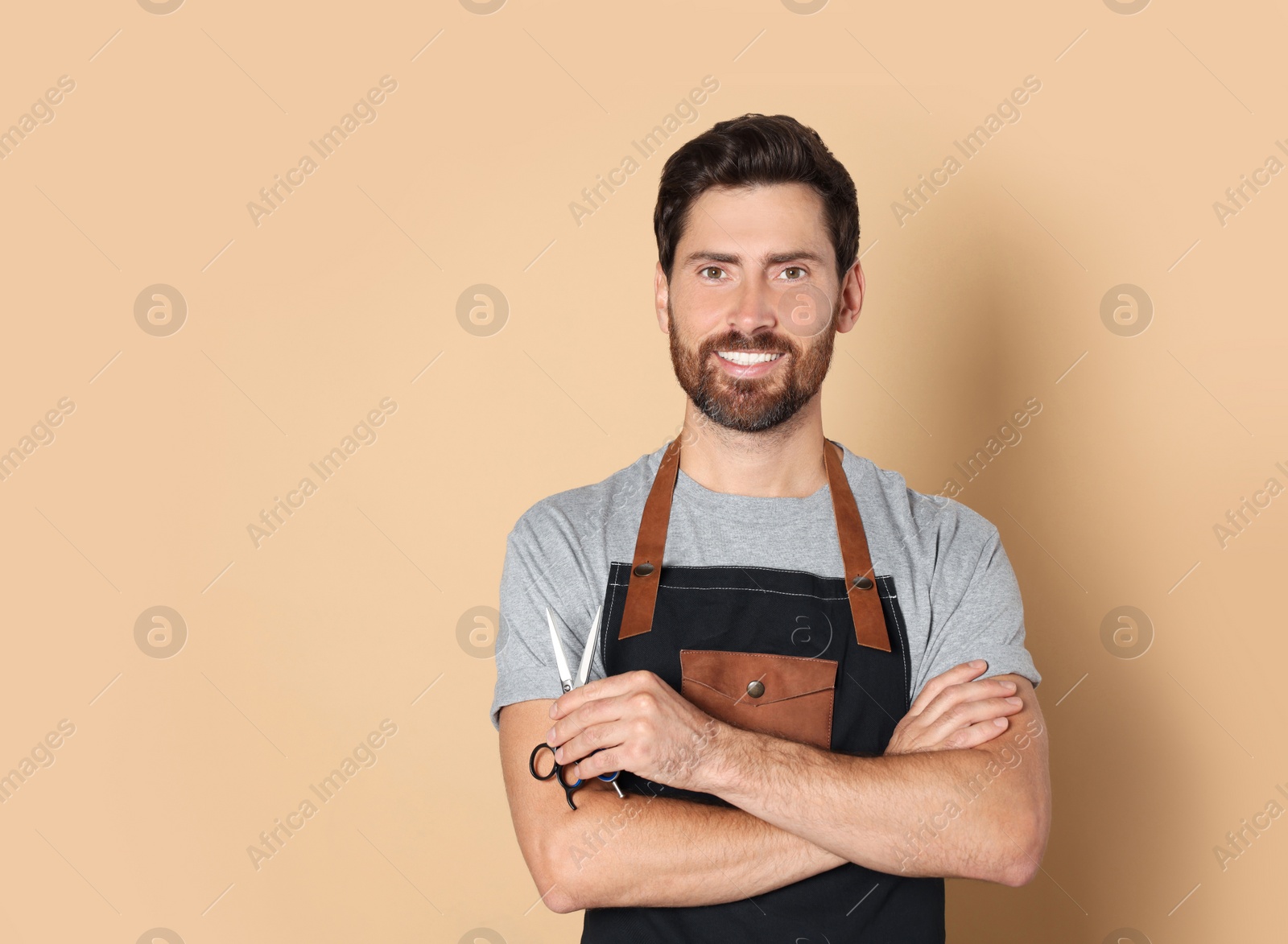 Photo of Smiling hairdresser in apron holding scissors on light brown background, space for text