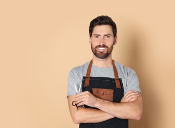 Smiling hairdresser in apron holding scissors on light brown background, space for text