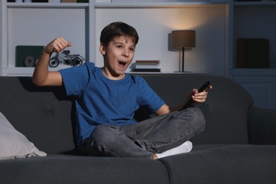 Emotional boy watching TV and holding remote control on sofa at home