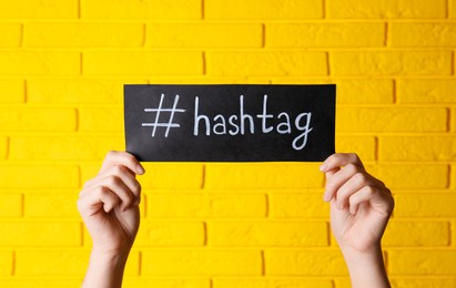 Woman holding black paper with word Hashtag and symbol against yellow brick wall, closeup