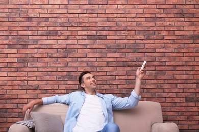 Young man operating air conditioner with remote control near brick wall at home