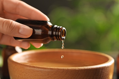 Photo of Woman pouring essential oil from glass bottle into bowl against blurred green background, closeup