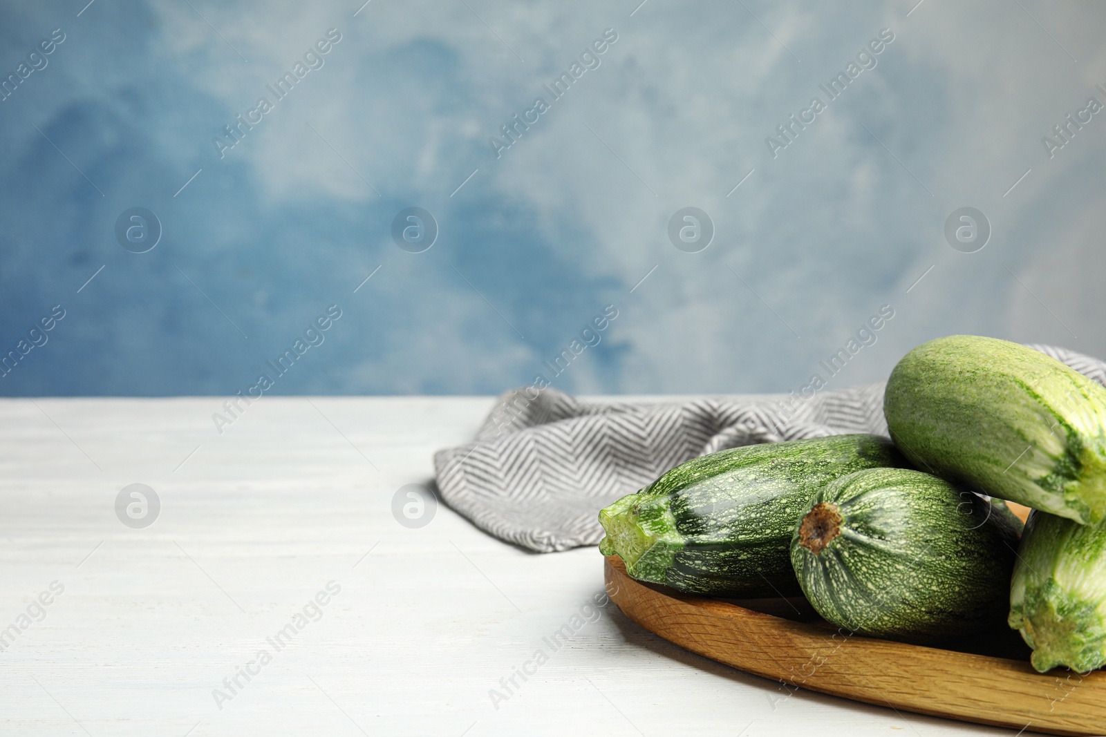 Photo of Wooden plate with fresh ripe green zucchinis on white table against blue background, space for text