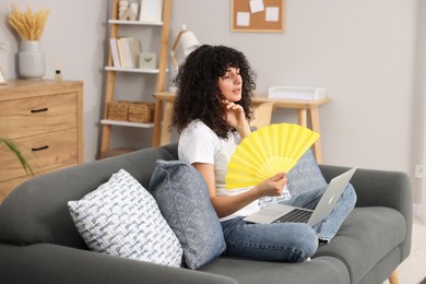 Young woman with laptop waving yellow hand fan to cool herself on sofa at home