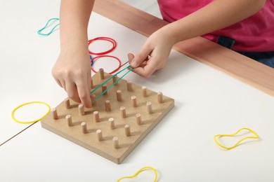 Photo of Motor skills development. Girl playing with geoboard and rubber bands at white table, closeup