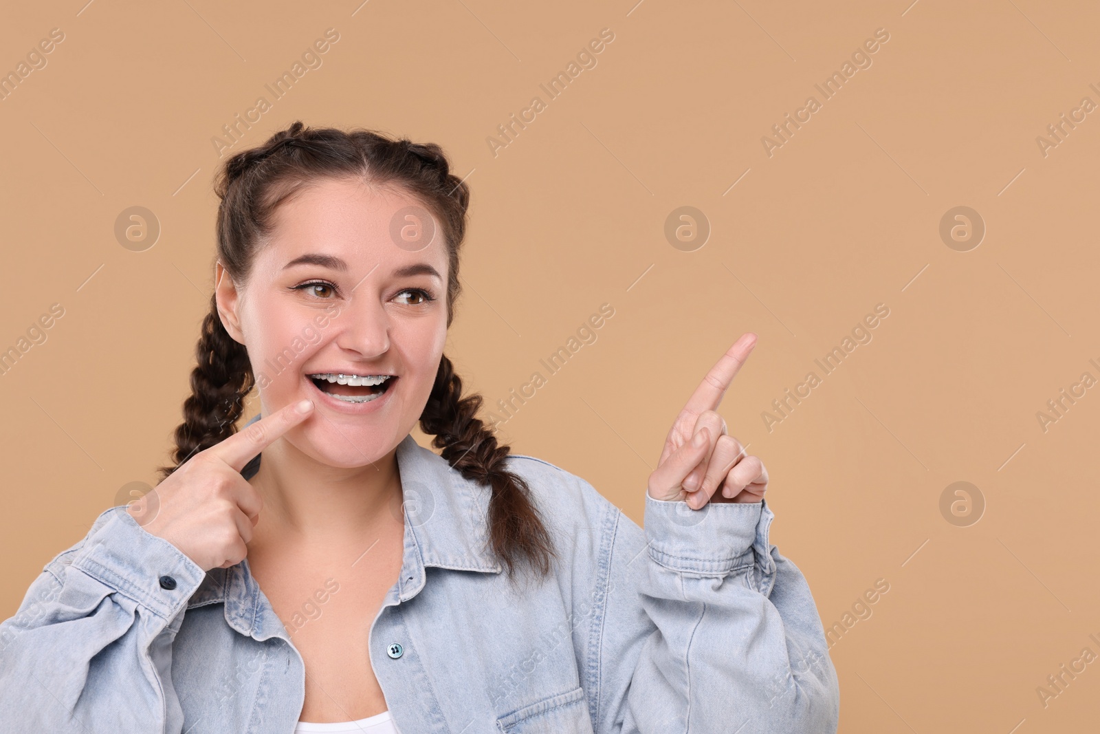 Photo of Smiling woman with braces pointing at something on beige background. Space for text