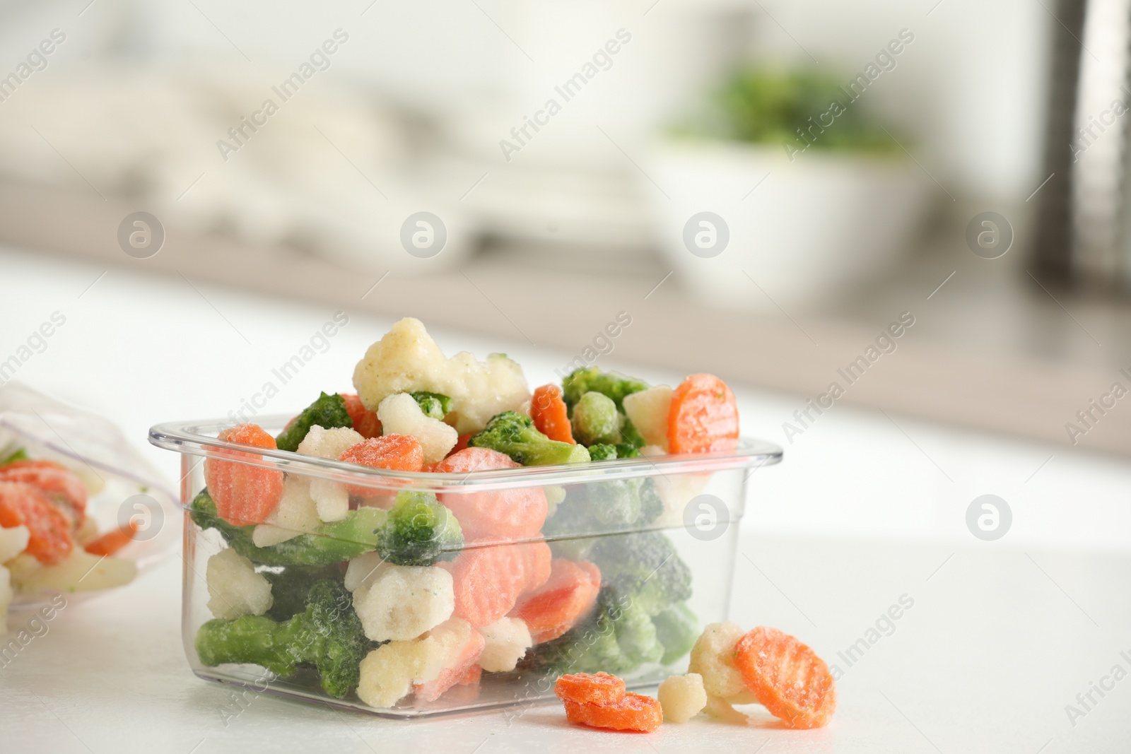 Photo of Mix of different frozen vegetables in plastic container on white textured countertop in kitchen, closeup. Space for text