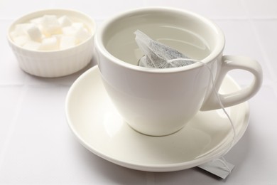 Photo of Tea bag in cup with hot water and bowl with sugar on white tiled table, closeup