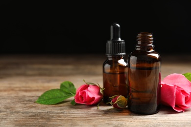 Photo of Bottles of rose essential oil and flowers on wooden table against black background, space for text