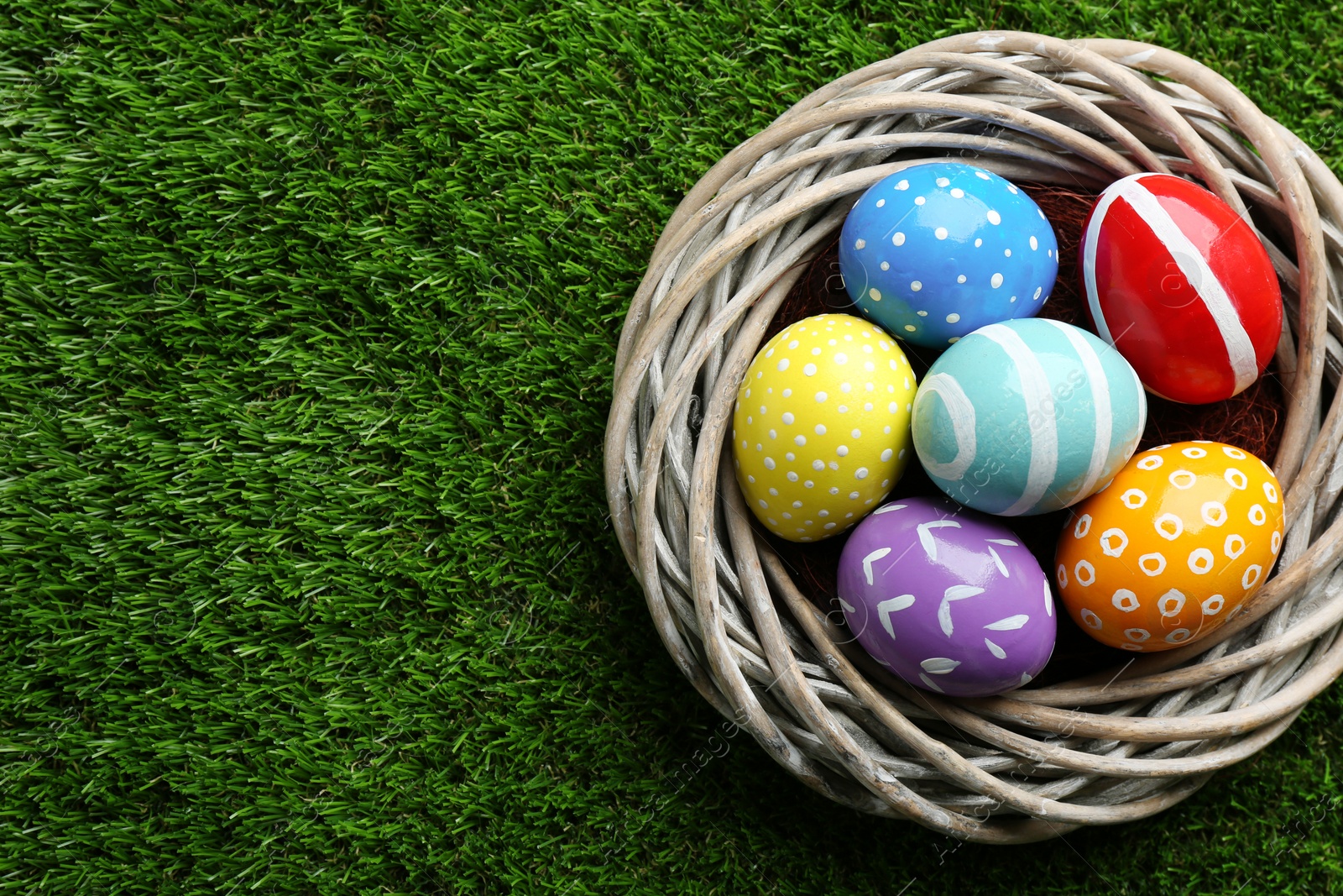 Photo of Wicker nest with painted Easter eggs on green grass, top view. Space for text