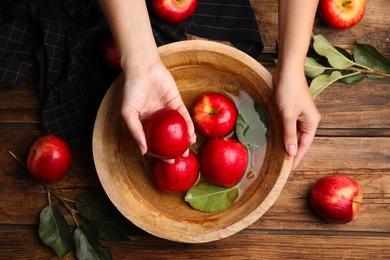 Woman washing ripe red apples in bowl of water at wooden table, top view