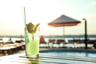 Glass of fresh summer cocktail on wooden table near swimming pool outdoors at sunset. Space for text