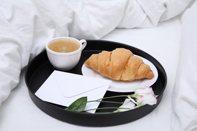 Photo of Tray with tasty croissant, cup of coffee and flowers on white bed