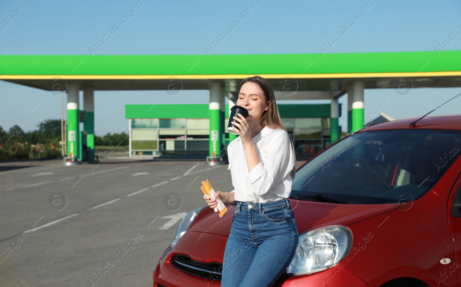 Photo of Beautiful young woman with hot dog drinking coffee near car at gas station