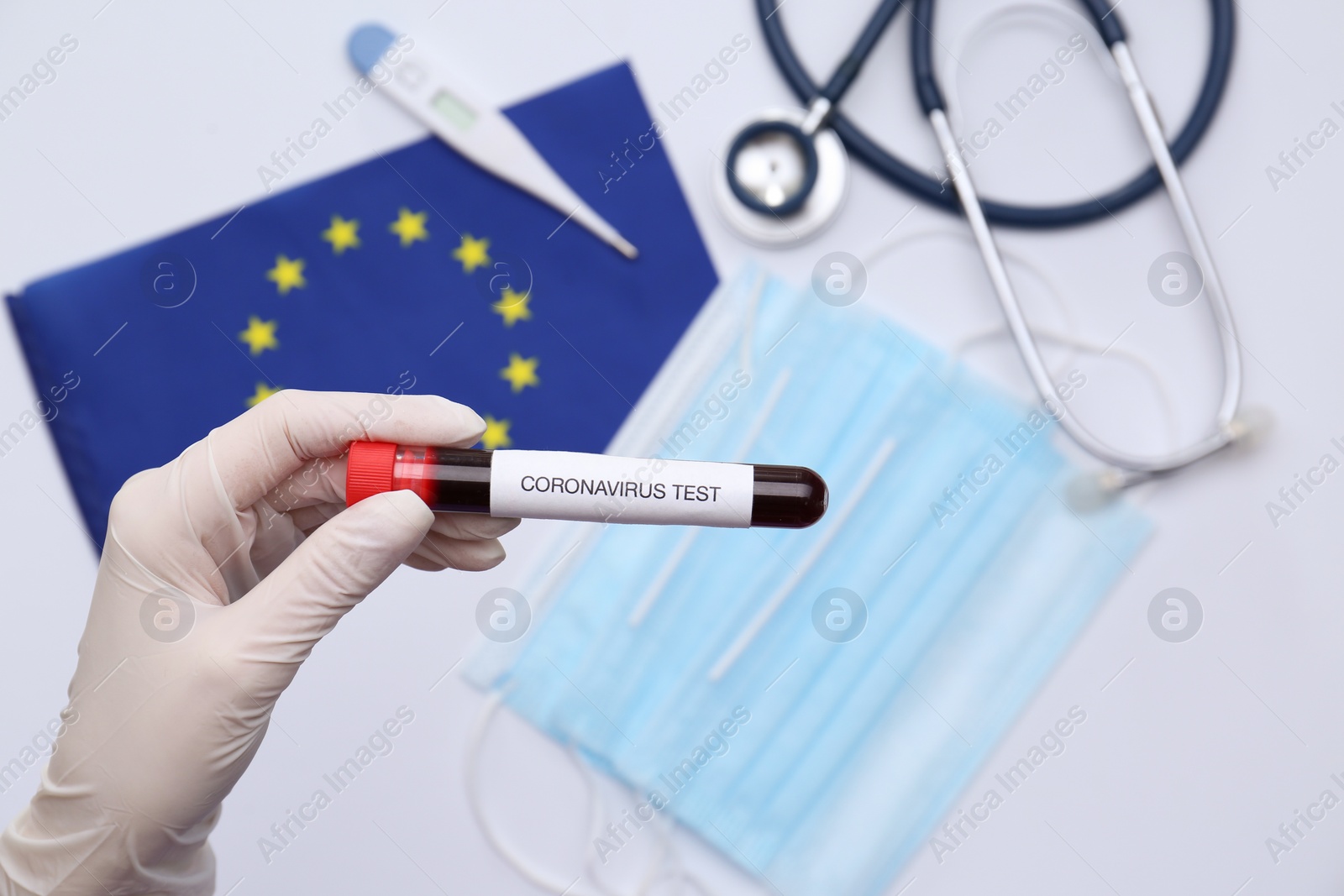 Photo of Doctor holding sample tube with label Coronavirus Test above medical items and European Union flag, closeup