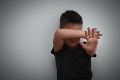 Photo of Scared little boy closing eyes with hand near white wall, focus on hand. Child in danger