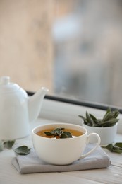 Photo of Cup of sage tea and green leaves on white wooden windowsill