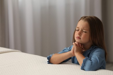 Cute little girl with hands clasped together saying bedtime prayer in bedroom. Space for text