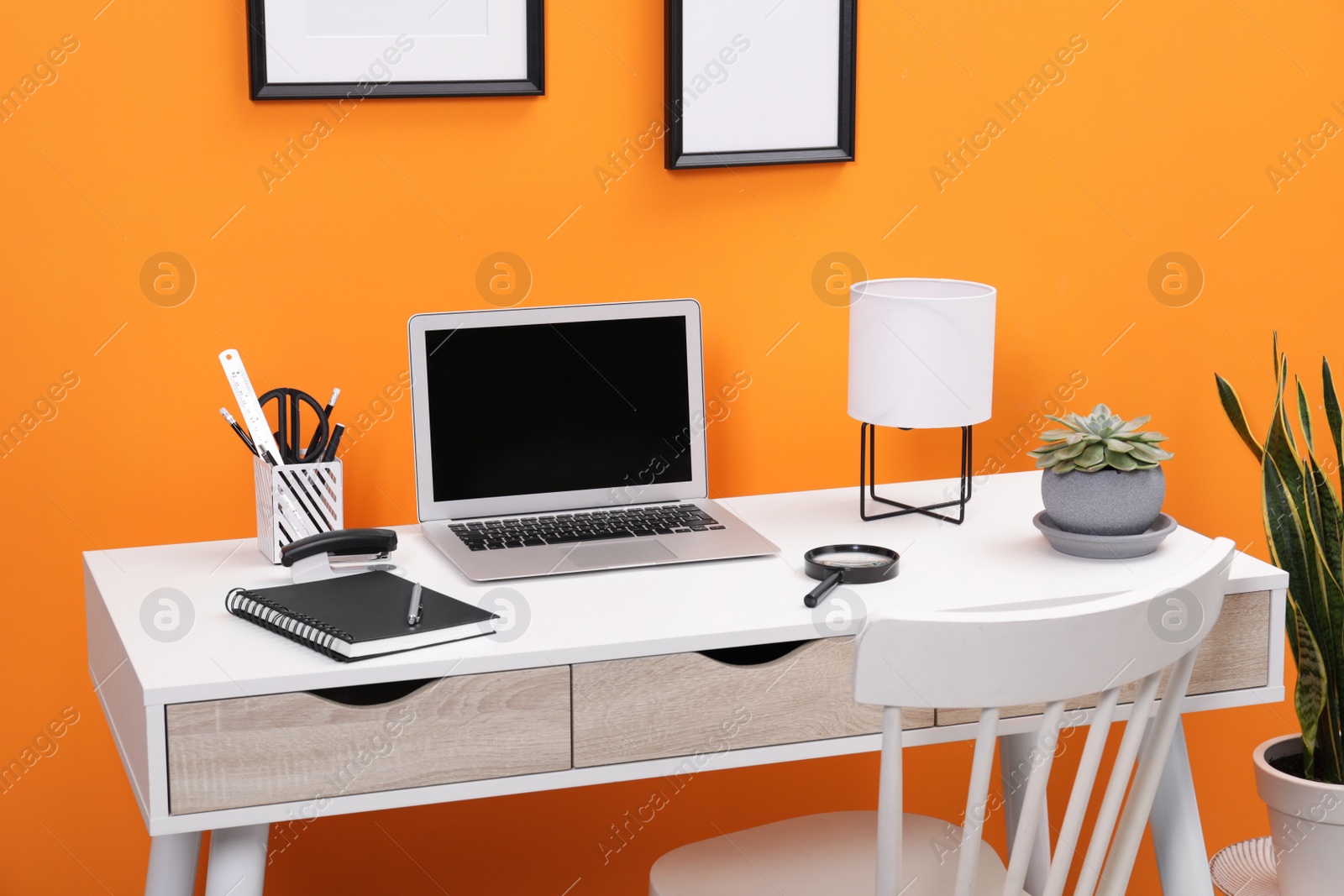 Photo of Workplace with laptop, stationery on desk and chair near orange wall. Home office