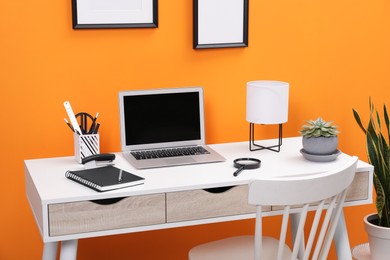 Workplace with laptop, stationery on desk and chair near orange wall. Home office