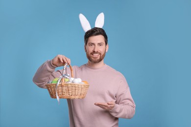 Photo of Portrait of happy man in cute bunny ears headband holding wicker basket with Easter eggs on light blue background