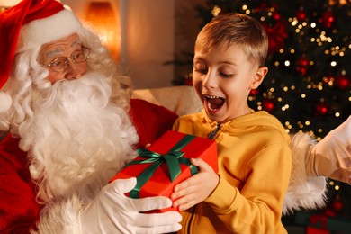 Surprised little boy taking gift from Santa Claus in room with Christmas tree