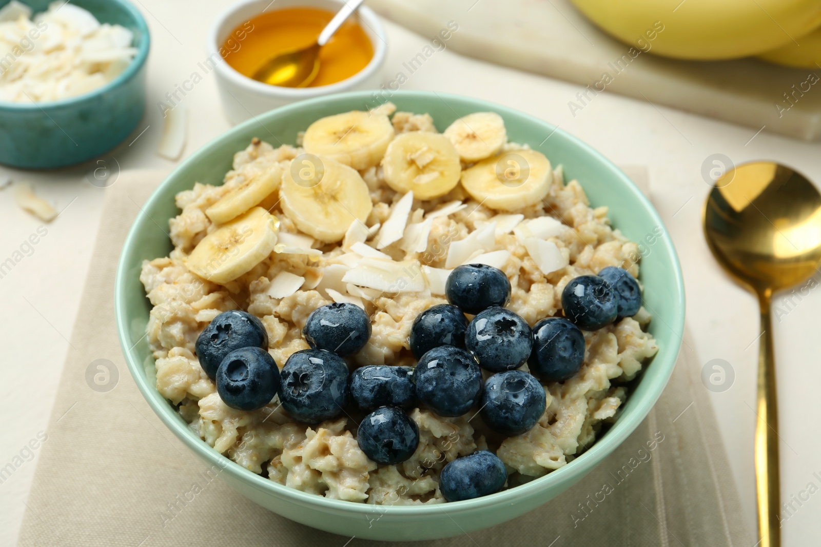 Photo of Tasty oatmeal with banana, blueberries, coconut flakes and honey served in bowl on beige table