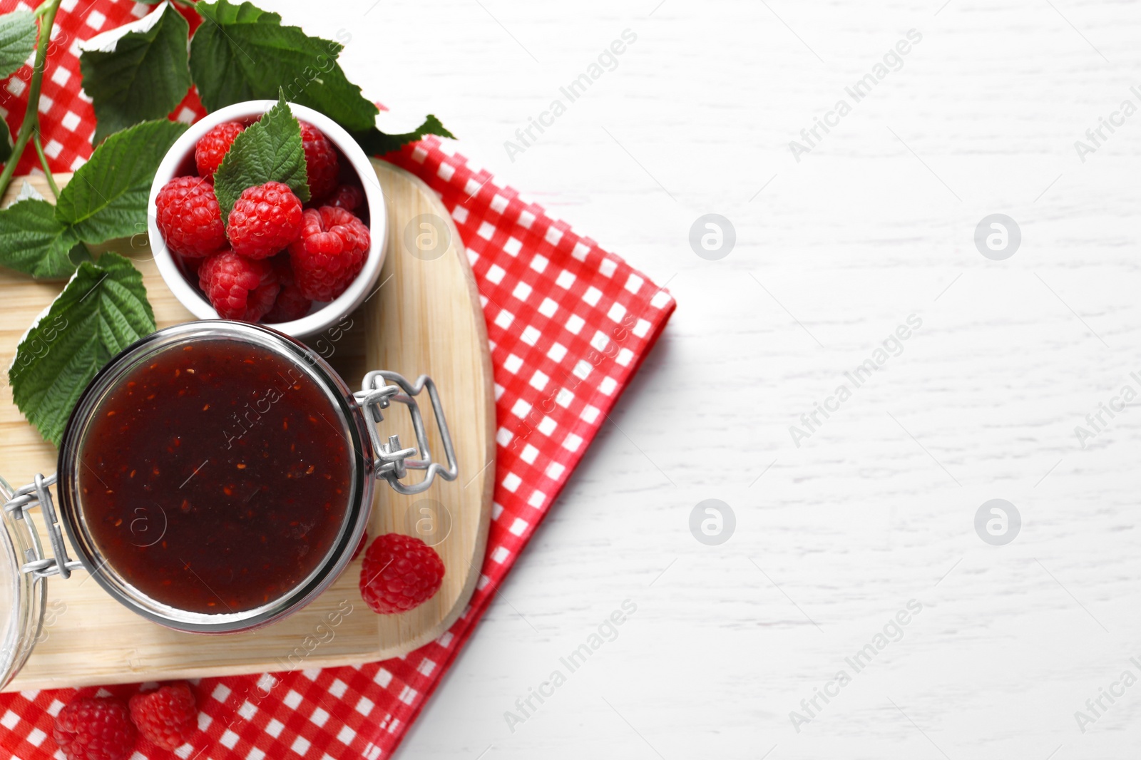 Photo of Jar of delicious raspberry jam, fresh berries and green leaves on white wooden table, top view. Space for text