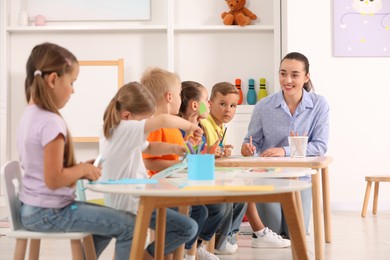 Photo of Nursery teacher with group of cute little children drawing and cutting paper at desks in kindergarten. Playtime activities