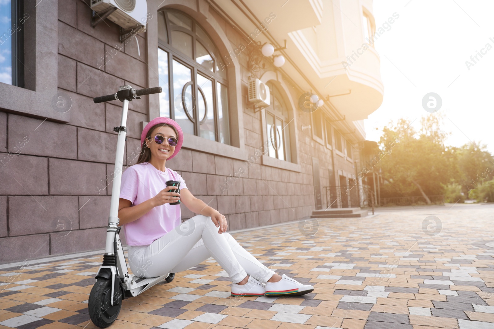 Photo of Young woman with cup of coffee sitting on electric kick scooter outdoors