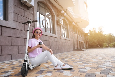 Young woman with cup of coffee sitting on electric kick scooter outdoors